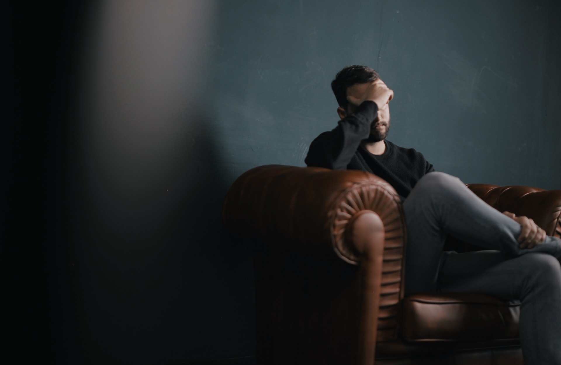Ein Mann sitzt auf einem Sofa in einem abgedunkelten Raum und hält sich die Hand an die Stirn. Symbolbild für Depression